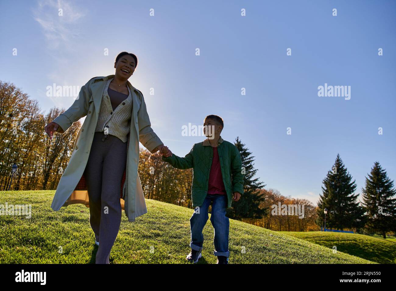 joyeux mère afro-américaine et enfant tenant la main, marchant sur la colline, tenues d'automne Banque D'Images