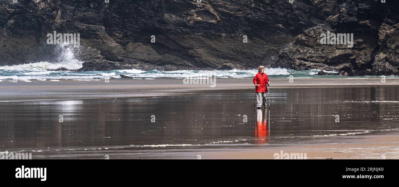 Une image panoramique d'une femme mature portant un manteau rouge vif et utilisant des bâtons de marche marchant des bâtons de randonnée marchant sur le rivage à la plage de Mawgan Porth à Cornw Banque D'Images