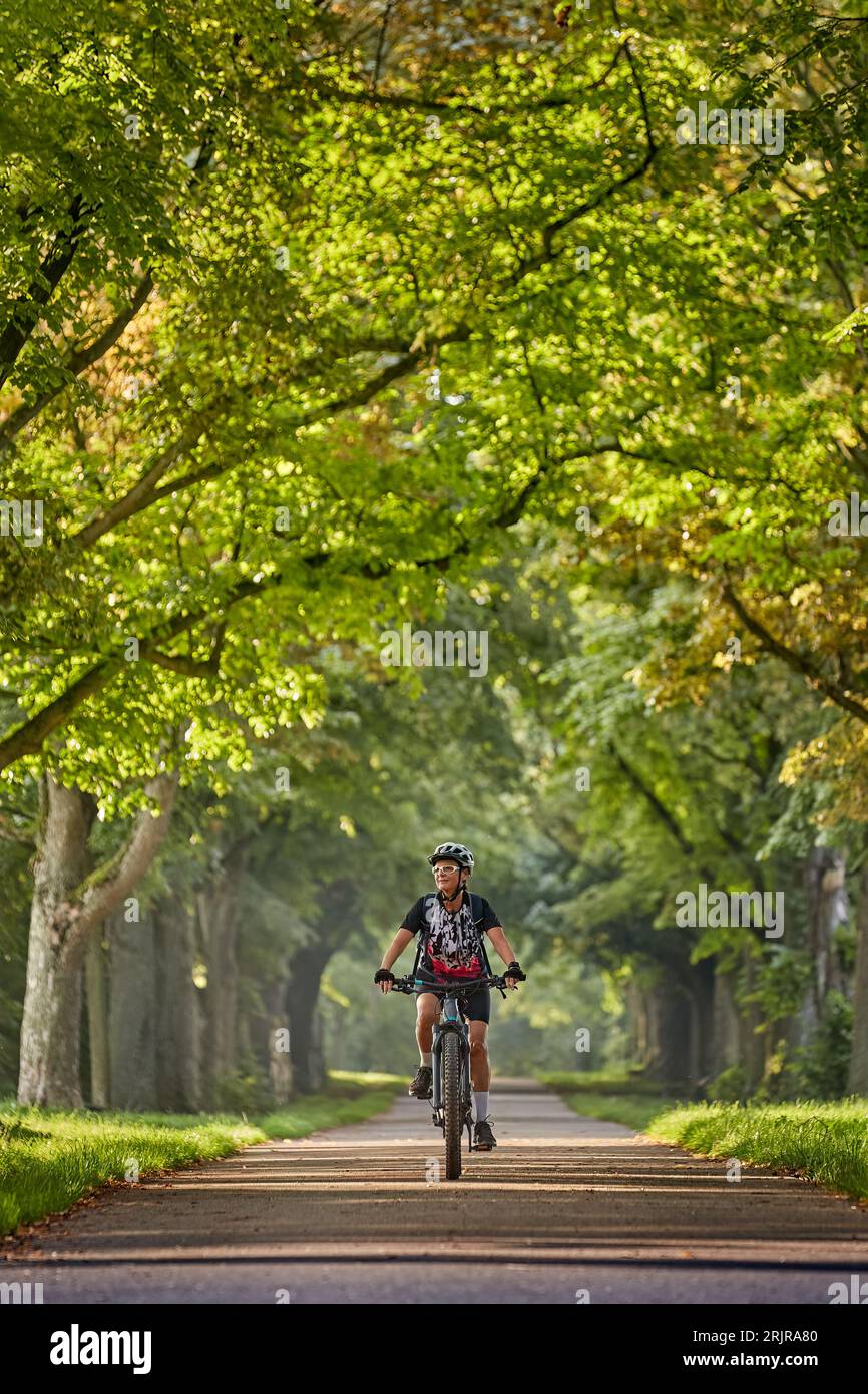Sénior femme sénior attrayante à vélo avec son vélo de montagne électrique dans une belle avenue de chêne et châtaignier à Ludwigsburg, Baden-Wuerttemberg, GE Banque D'Images