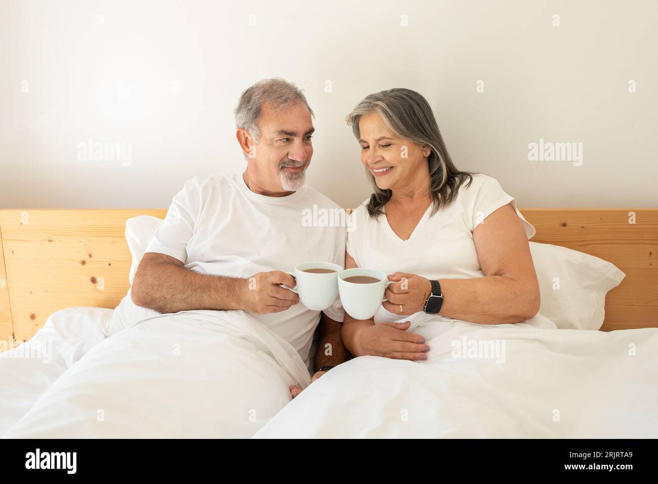 Souriant vieux mari et femme caucasiens se trouve au lit, acclament avec des tasses de café, profitez du bonjour Banque D'Images