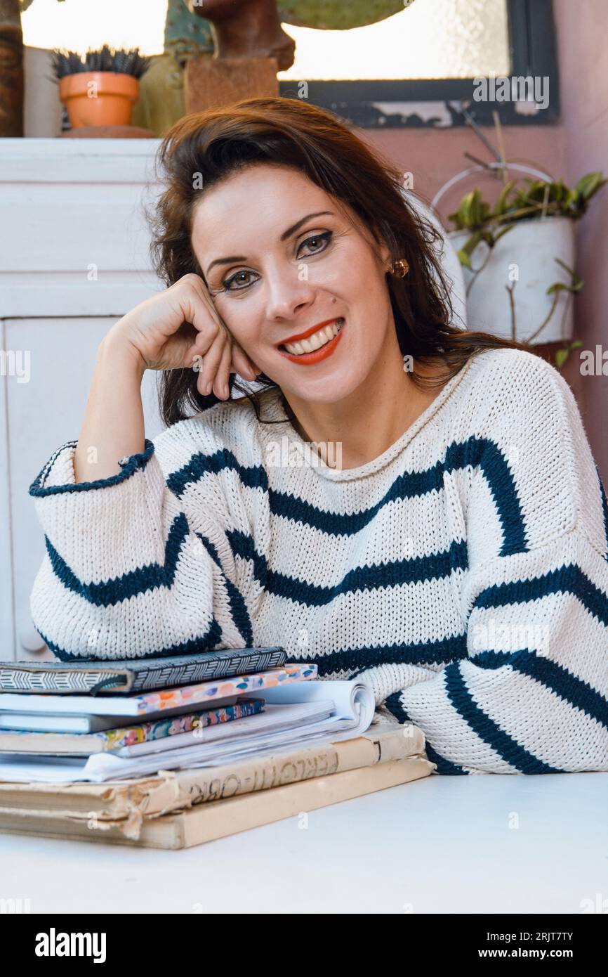 portrait de femme étudiante adulte caucasienne française portant un chandail rayé bleu heureux assis souriant et regardant la caméra appuyée sur la table avec des livres, Banque D'Images