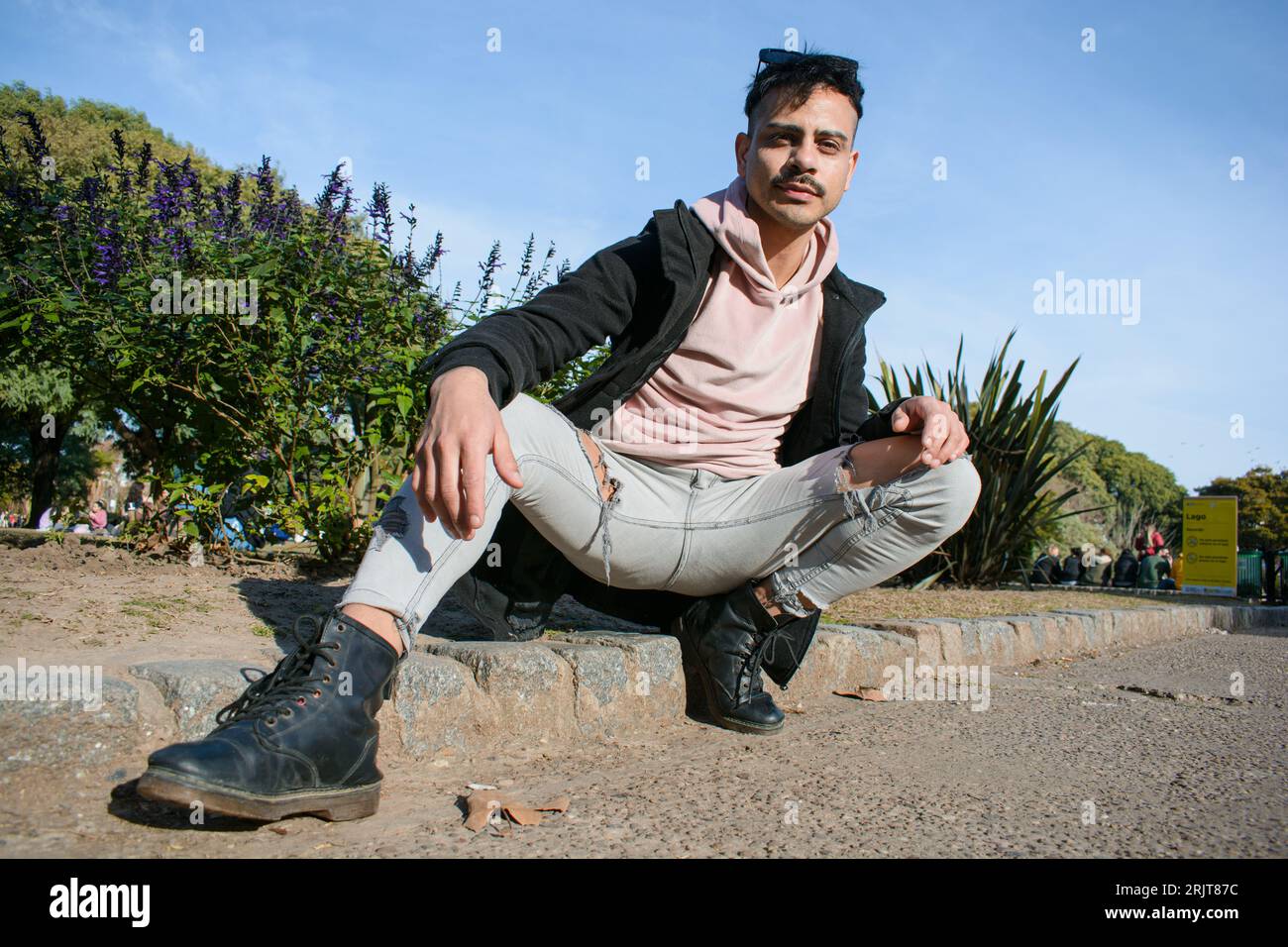 portrait de beau jeune homme homosexuel latino d'origine argentine, avec moustache et cheveux courts, assis avec les jambes ouvertes à l'extérieur en public pair Banque D'Images