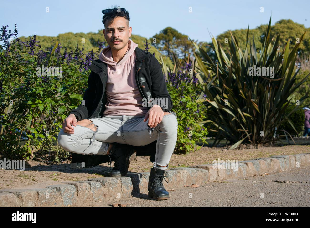 jeune homme latino homosexuel à la mode avec moustache et cheveux courts, porte un pull rose et une veste noire, accroupi posant en regardant la caméra dans le parc Banque D'Images