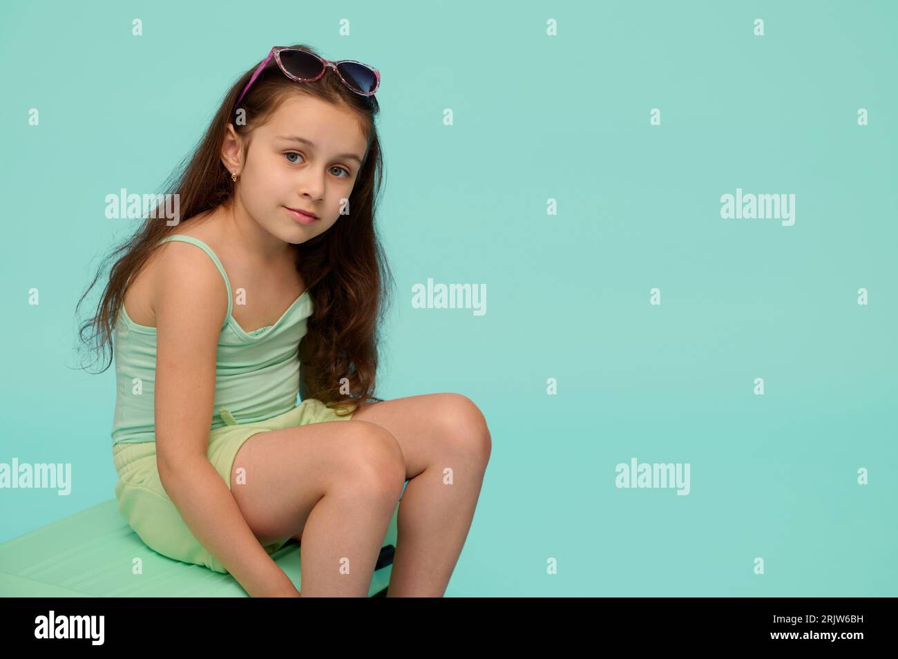 Belle enfant fille, voyageur touriste, passager, en bleu turquoise tendance dans les vêtements d'été, assis sur une valise en plastique, souriant mignon regardant c Banque D'Images