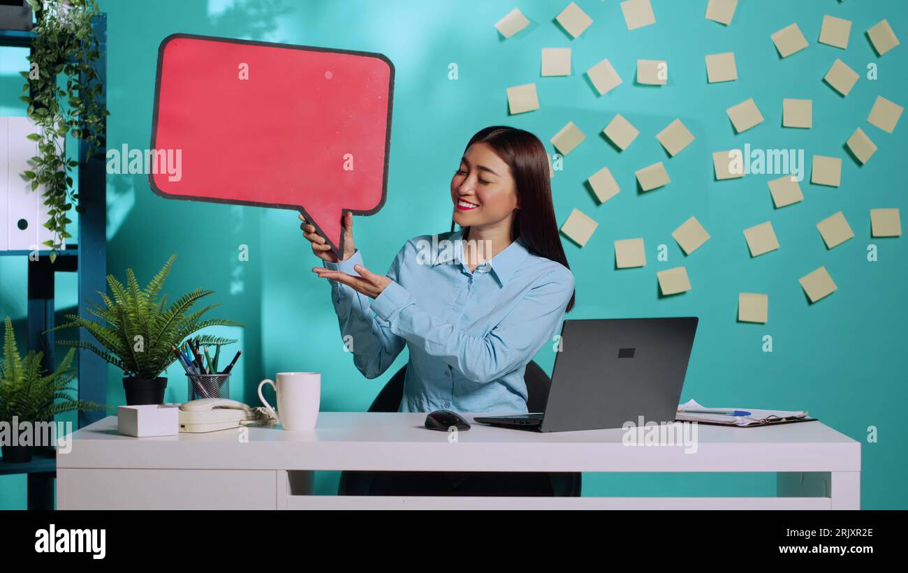 Heureuse joyeuse femme d'affaires asiatique tenant le signe rouge de bulle de discours de l'espace vide pour le texte et montrant le geste de main ok. Employé souriant présentant du carton à bulles de pensée dans un lieu de travail coloré Banque D'Images