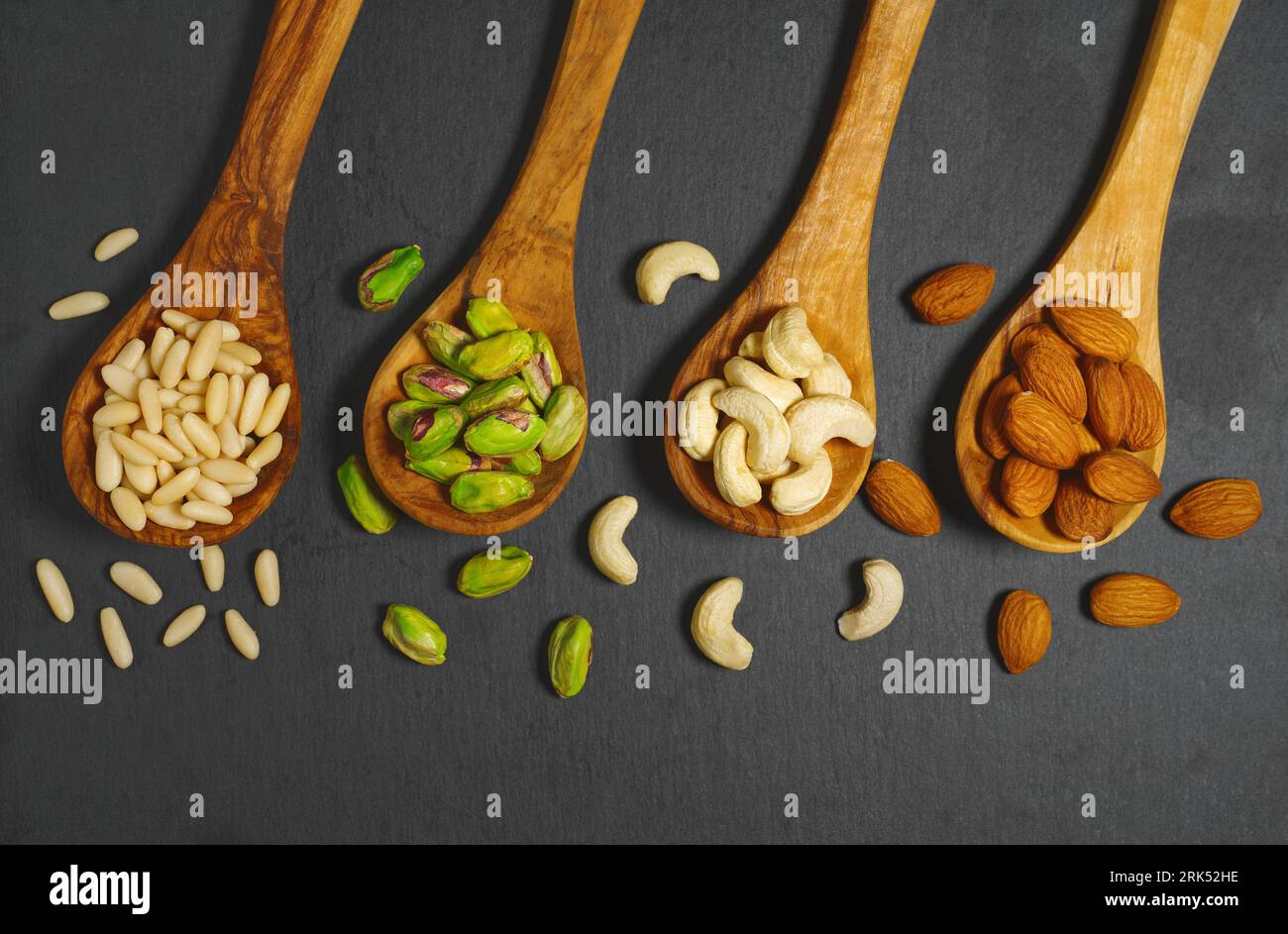 Les noix se mélangent avec des pignons de pin, des pistaches, des noix de cajou et des amandes dans des cuillères en bois vue de dessus gros plan sur fond gris foncé. Banque D'Images