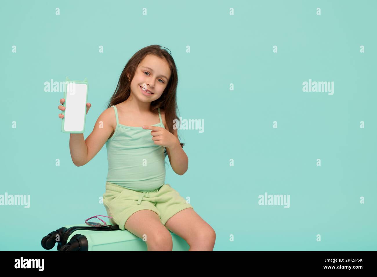 Adorable petite fille joyeuse assise sur sa valise, souriant et montrant à la caméra un smartphone moderne avec maquette écran numérique blanc, isolé ov Banque D'Images