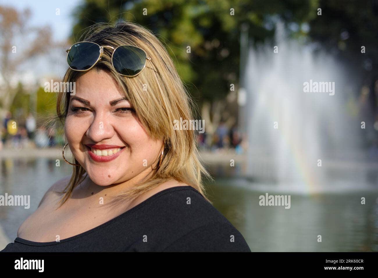 gros plan portrait de jeune femme blonde plus taille latina, debout dans le parc public heureux portant des lunettes de soleil, posant souriant et regardant la caméra, avec Banque D'Images