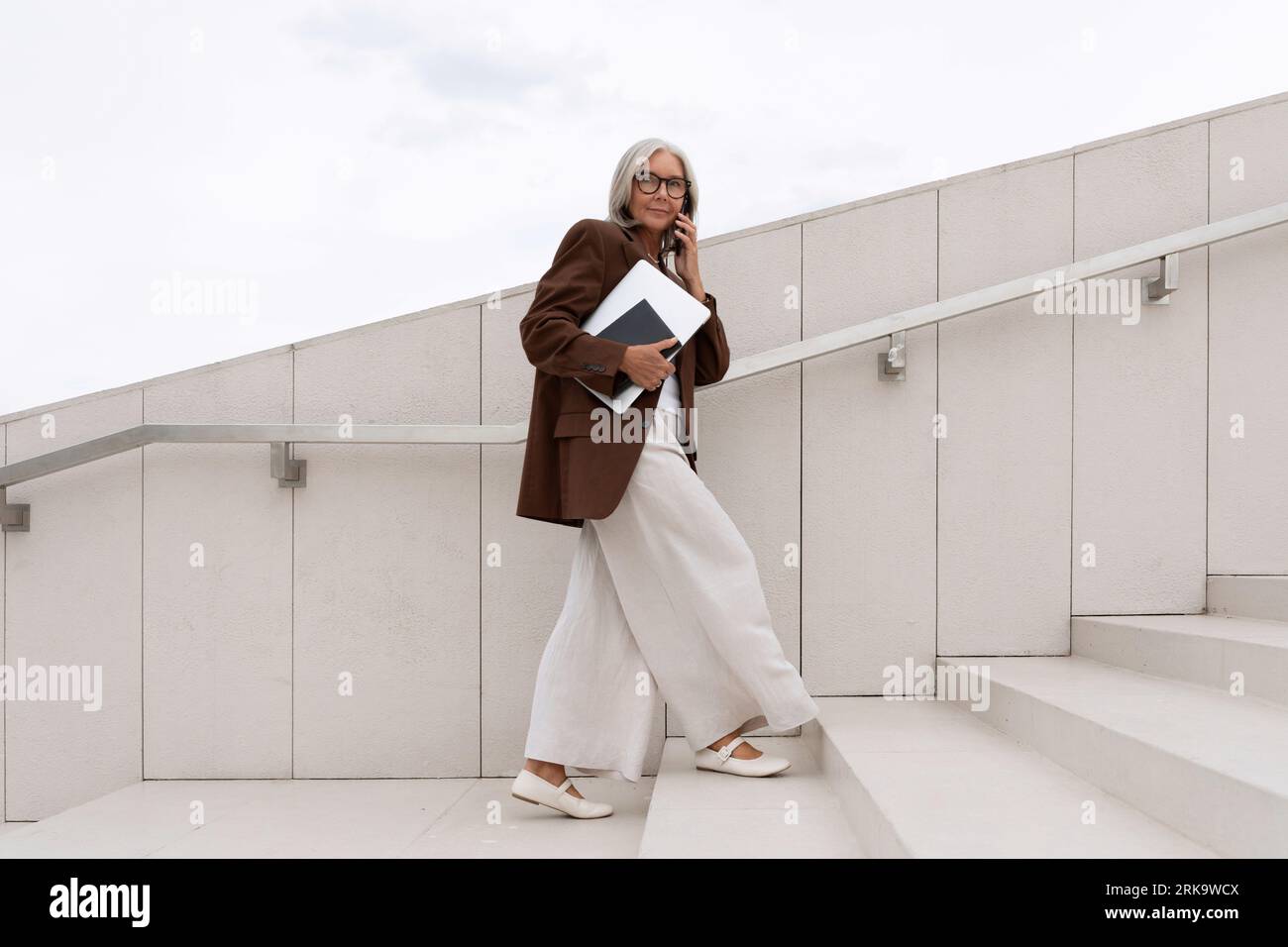 Confiante vieille dame mature avec les cheveux gris portant des lunettes portant une veste marron va à la réunion d'affaires sur les escaliers Banque D'Images