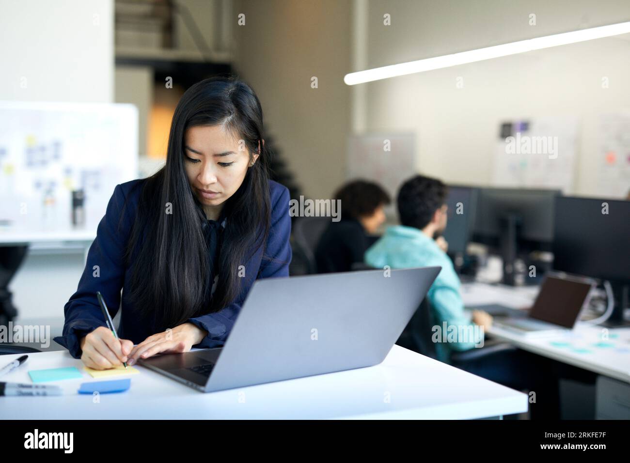 Femme d'affaires écrivant sur la note adhésive tandis que des collègues travaillant en arrière-plan au bureau Banque D'Images