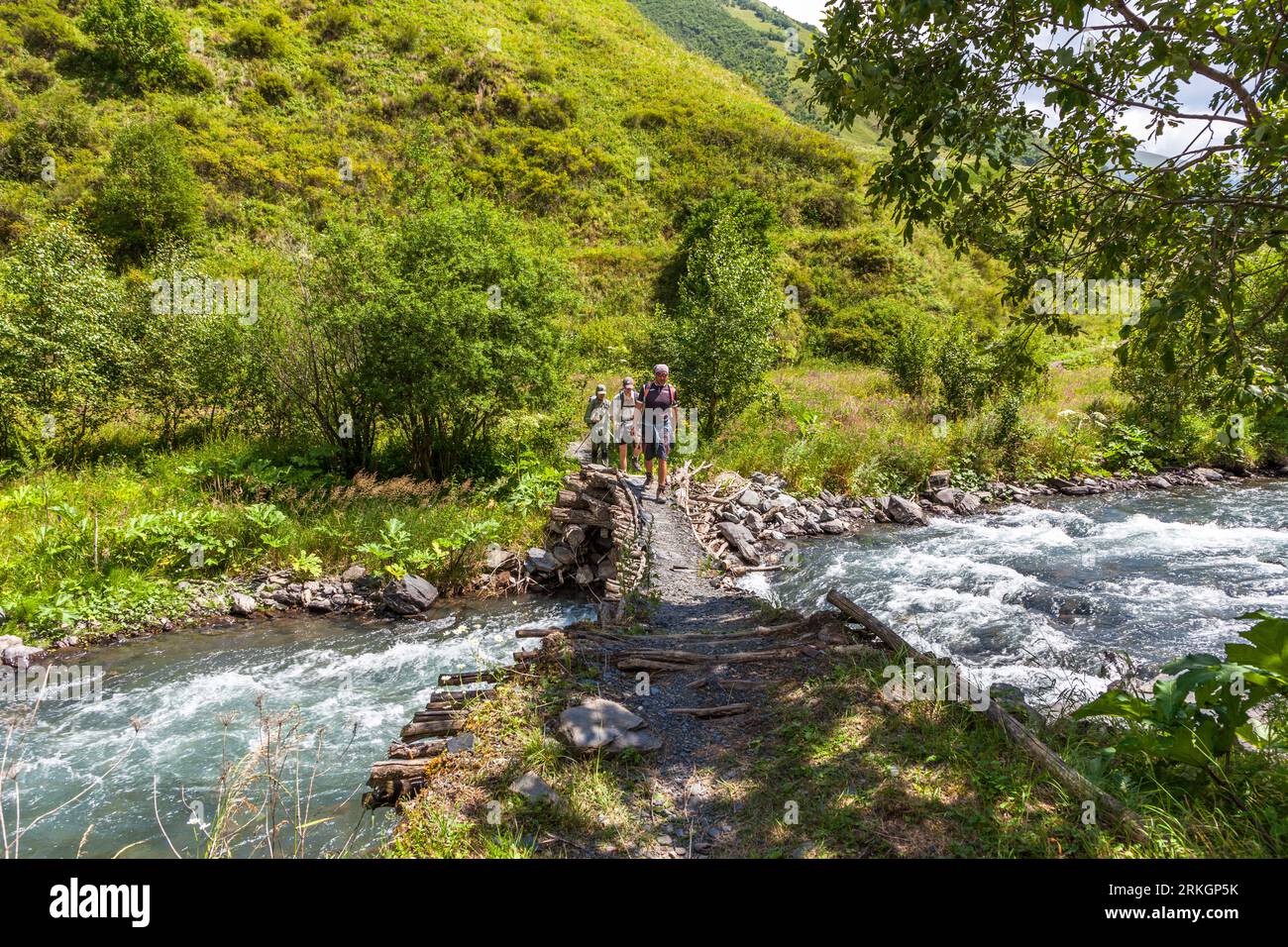 Trois randonneurs traversent une passerelle de troncs d'arbres et de pierres sur une rivière de montagne en file indienne près du village d'Ardoti dans la municipalité de Dusheti, région de Mtskheta-Mtianeti, Géorgie. L'eau mousseuse déferle des deux côtés de la passerelle. Des buissons et des arbres poussent sur la rive. Une pente verdoyante s'élève derrière eux. La randonnée avec des chevaux de bât a parcouru près de 120 kilomètres et plus de 5 000 mètres de dénivelé. Le village largement abandonné se trouve sur une colline au-dessus de la vallée d'Andaki, à environ 5 kilomètres au sud-ouest de Mutso. Khevsouréti (géorgien ხევსურეთი) est une région historique sur les pentes du Grand Banque D'Images