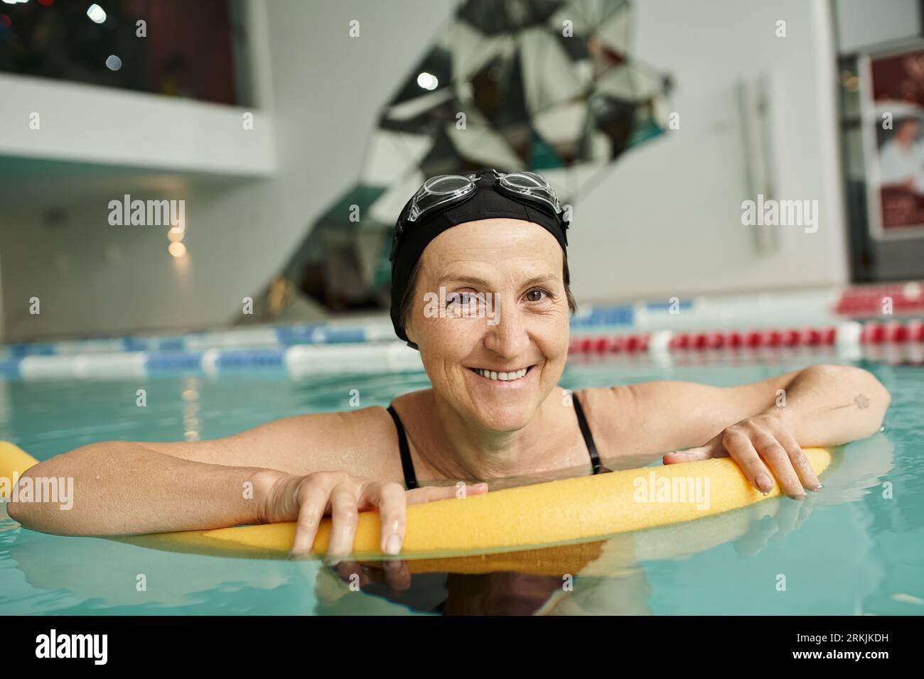 heureuse femme d'âge moyen en bonnet de bain et lunettes nageant avec nouilles de piscine, mode de vie sain, sport Banque D'Images