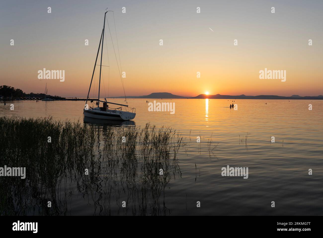 Lac Balaton au coucher du soleil avec les gens et la silhouette de voilier et les montagnes de la partie nord en arrière-plan. Banque D'Images