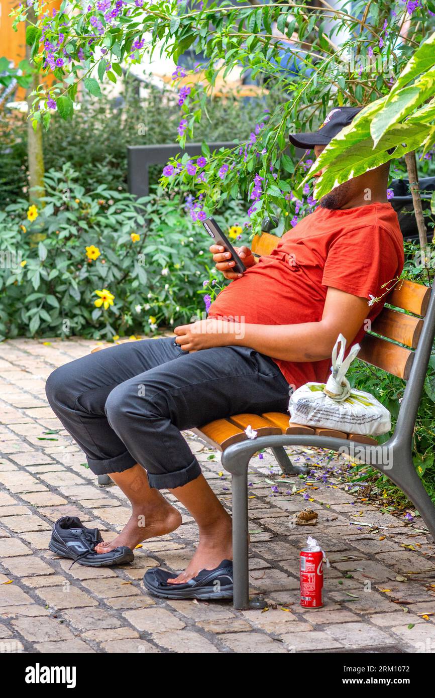 Homme noir assis sur un banc de parc avec le visage caché à l'aide d'un smartphone - Tours, Indre-et-Loire (37), France. Banque D'Images