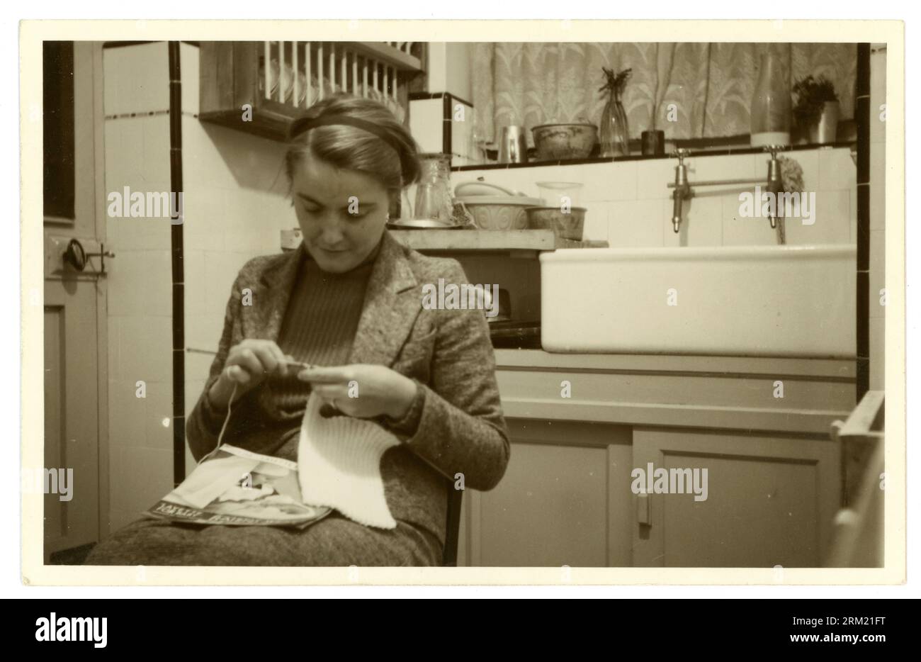 Photographie originale des années 1950 d'une jeune femme assise dans sa cuisine, portant un costume en tweed et un pull côtelé skinny dessous. Une femme qui travaille ou peut-être dans des fouilles d'étudiants. La jeune femme tricote, en utilisant un motif de laine, Royaume-Uni Banque D'Images