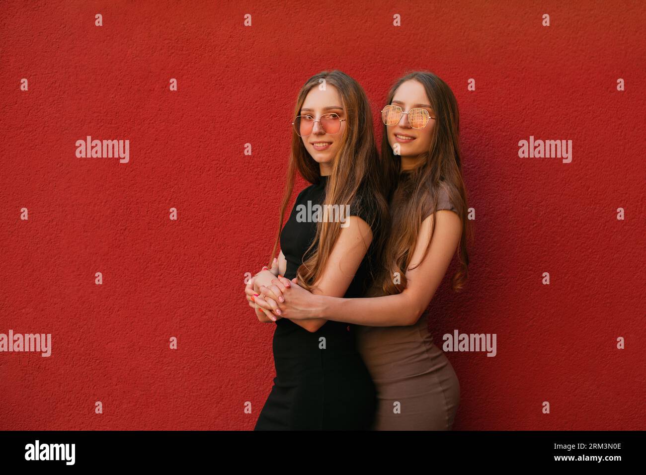 Heureux deux jeunes sœurs jumelles décontractées drôles avec des lunettes de soleil à la mode regardant la caméra debout sur un fond de mur rouge à l'extérieur jour d'été Banque D'Images