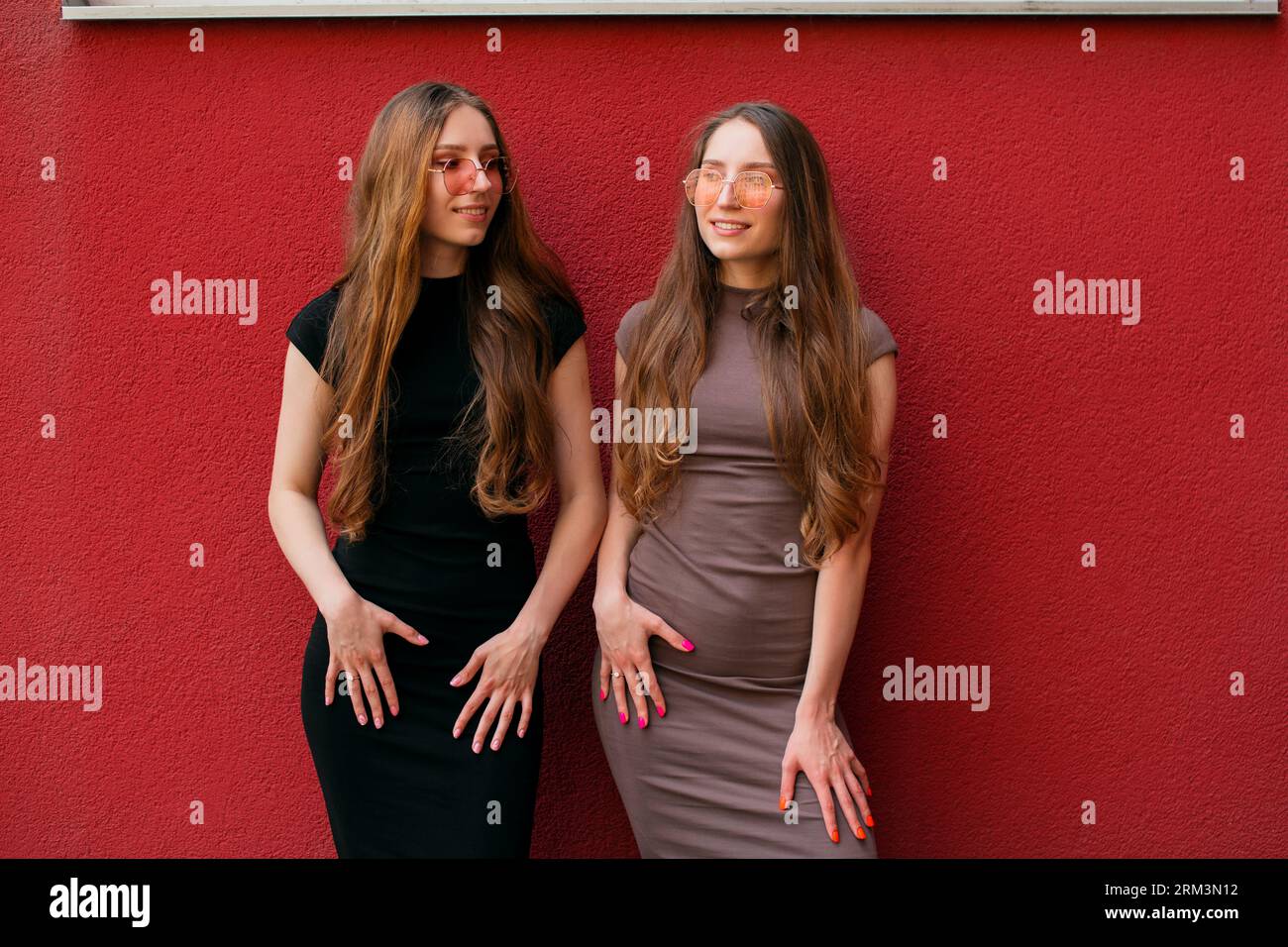 Heureux deux jeunes sœurs jumelles décontractées drôles avec des lunettes de soleil à la mode regardant la caméra debout sur un fond de mur rouge à l'extérieur jour d'été Banque D'Images