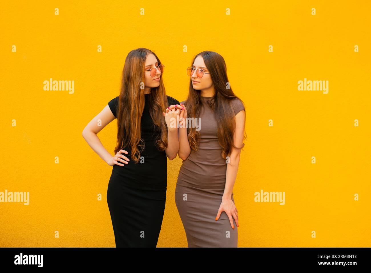Connexion entre drôle deux jeunes soeurs jumelles génération Z avec des lunettes de soleil fraîches tenant les mains regardant la caméra debout sur fond de mur jaune Banque D'Images