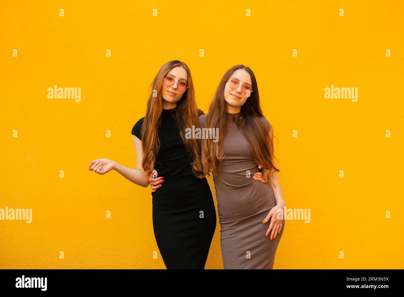 Connexion entre drôle deux jeunes soeurs jumelles génération Z avec des lunettes de soleil fraîches tenant les mains regardant la caméra debout sur fond de mur jaune Banque D'Images