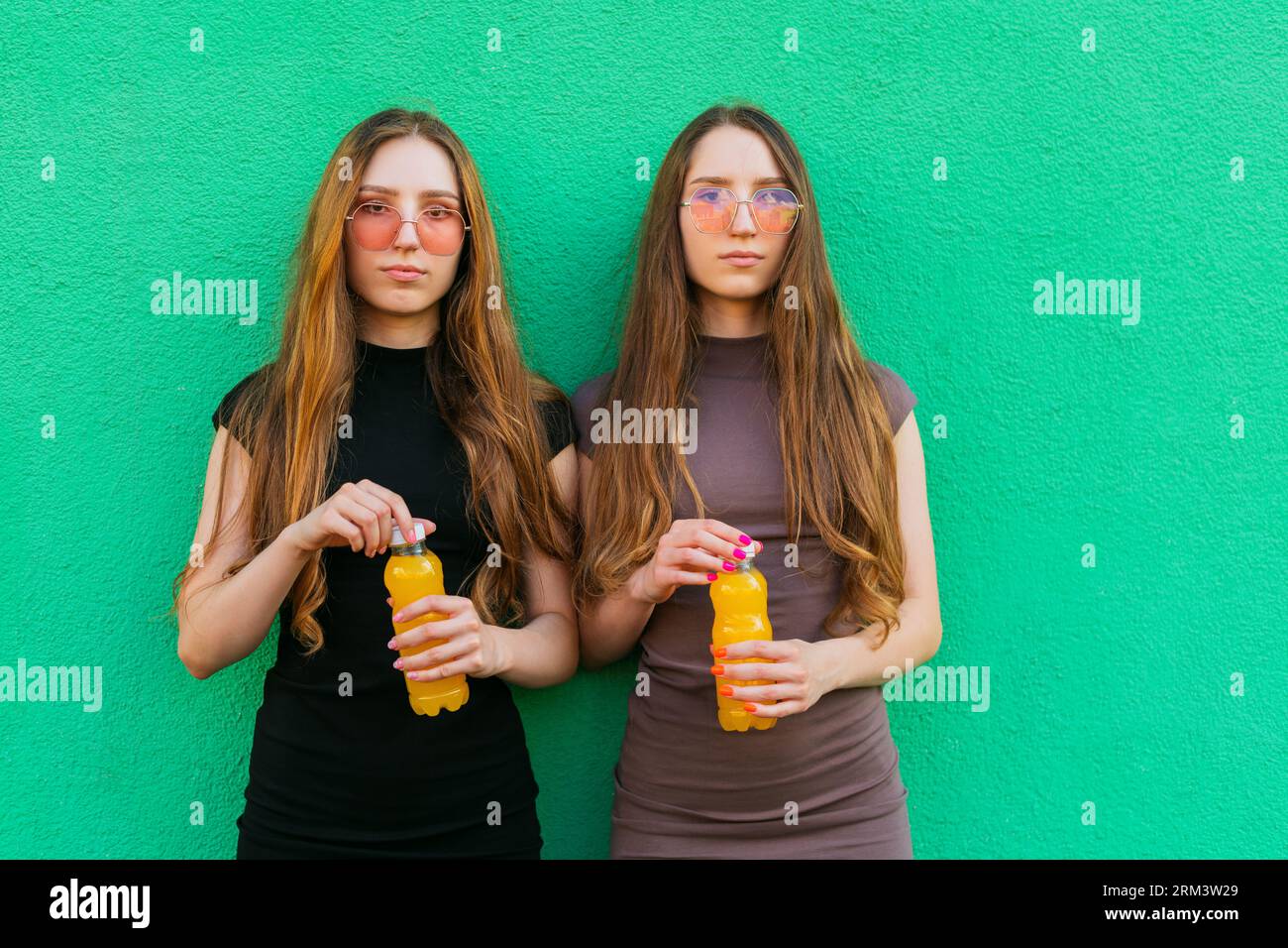 Jeunes sœurs jumelles mignonnes avec des lunettes de soleil cool tenant des bouteilles en plastique limonade jaune debout sur un fond de mur vert Banque D'Images
