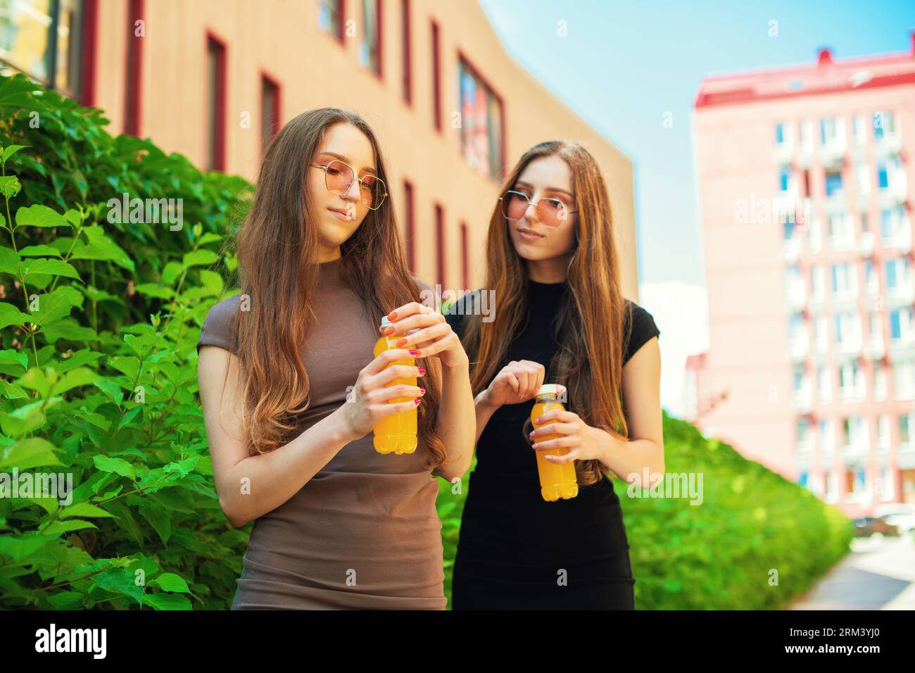 Jeunes sœurs jumelles mignonnes urbaines avec des lunettes de soleil fraîches ouvrant une bouteille orange de jus de citron debout dans le fond de la rue de la ville Banque D'Images
