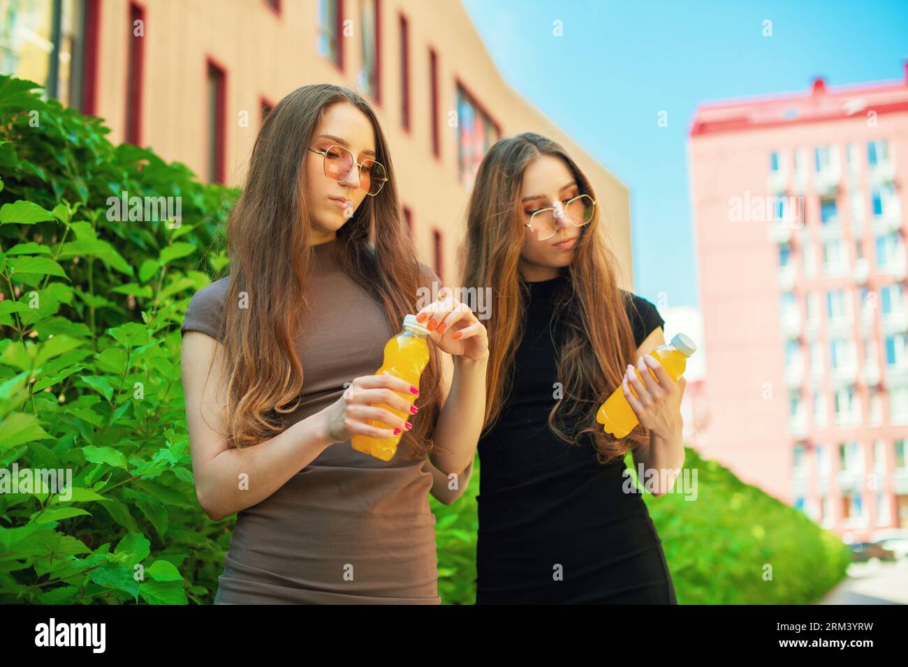 Jeunes sœurs jumelles mignonnes urbaines avec des lunettes de soleil fraîches ouvrant une bouteille orange de jus de citron debout dans le fond de la rue de la ville Banque D'Images
