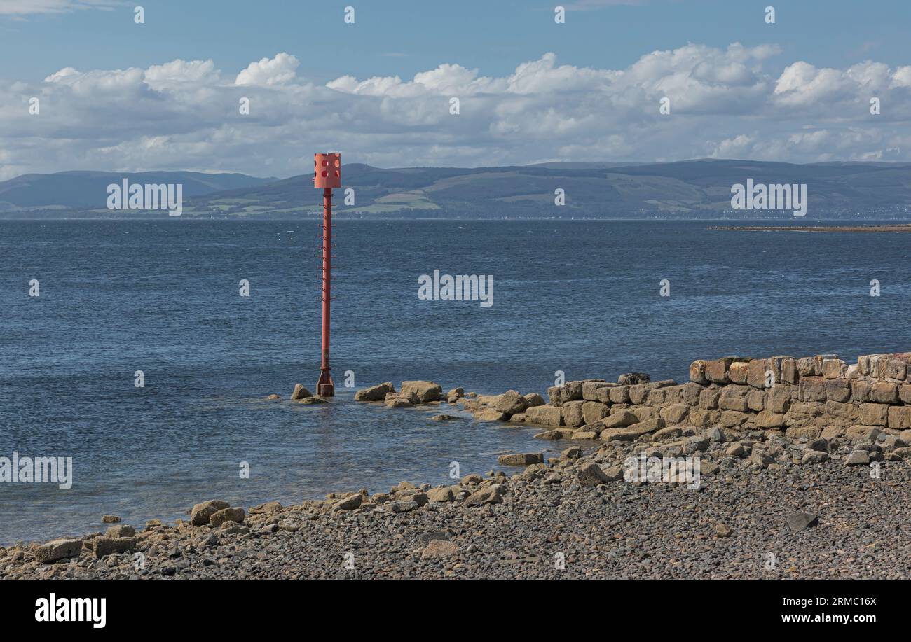 Balise peinte en orange sur le rivage à travers une plage pierreuse contre une mer bleue Banque D'Images