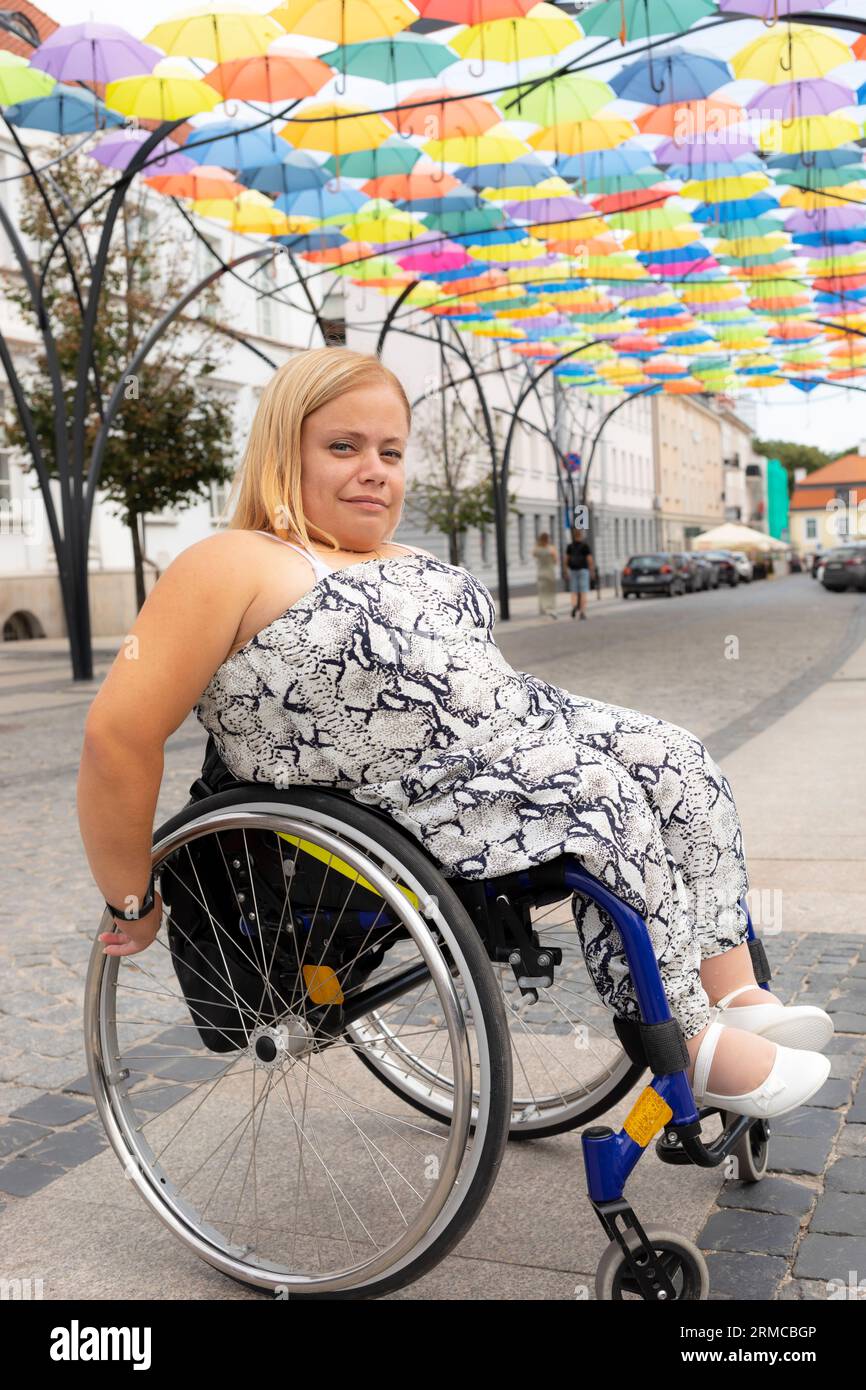 Jeune fille avec petite stature sur fauteuil roulant explore, se promène autour de la ville le jour de l'été. Femme adulte avec handicap physique appréciant en plein air et Banque D'Images