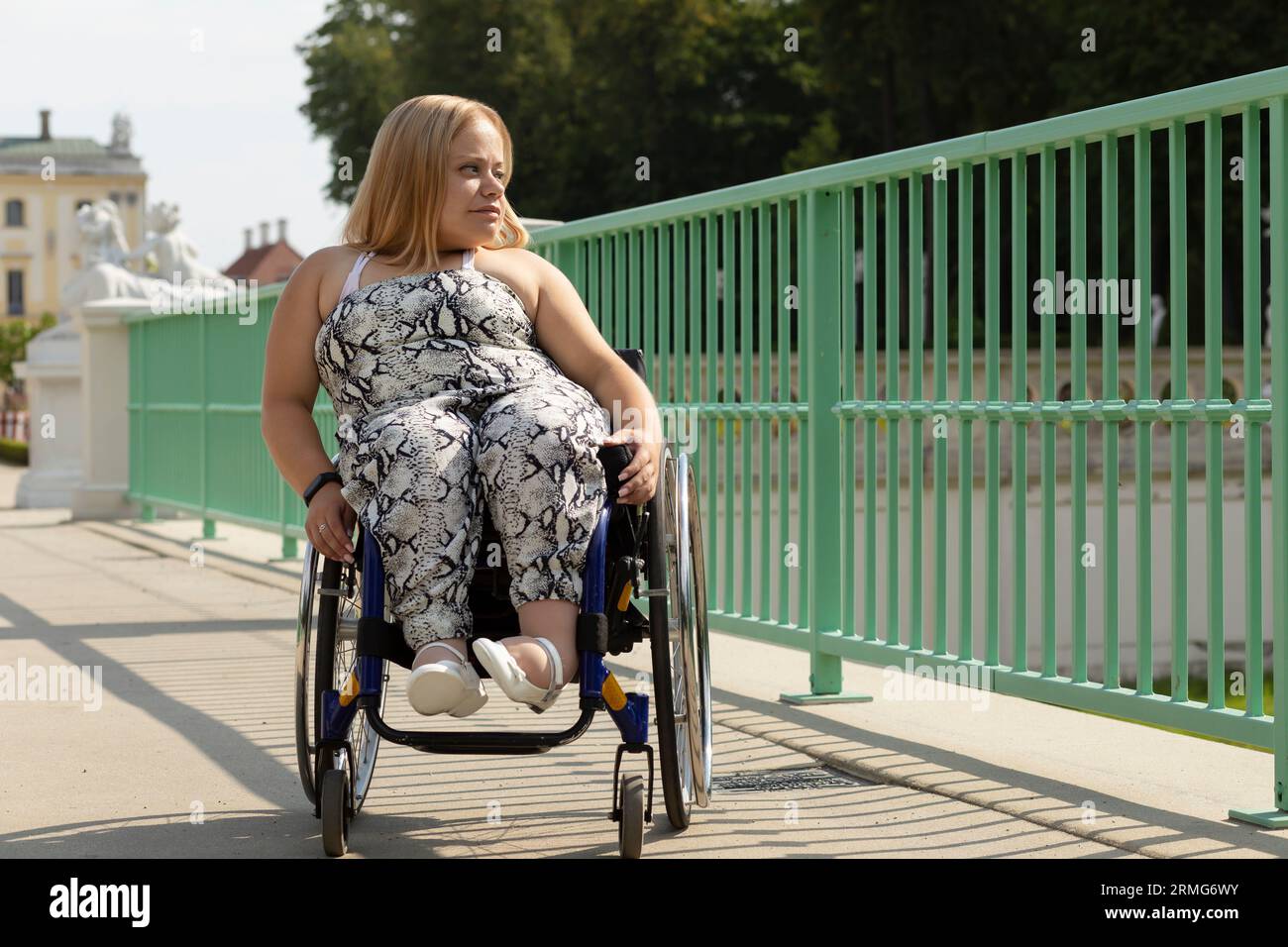 Belle jeune femme avec une courte hauteur marche sur le pont en fauteuil roulant dans la ville le jour d'été. Femme adulte handicapée. Copier l'espace pour le texte Banque D'Images