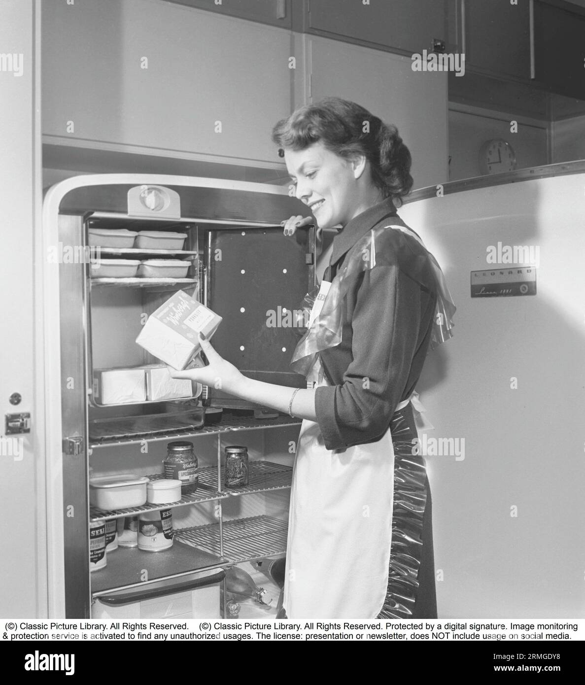 Dans la cuisine 1950s. Intérieur d'une cuisine et une jeune femme debout au réfrigérateur Leonard avec sa porte ouverte, montrant les boîtes et les bouteilles de différents aliments debout sur les étagères. Une armoire de congélation séparée est intégrée dans le réfrigérateur, une fonctionnalité assez nouvelle à ce moment. Elle porte un tablier en plastique. Elle est Haide Göransson, 1928-2008, mannequin et actrice. Suède 1950 Kristoffersson ref AU22-10 Banque D'Images