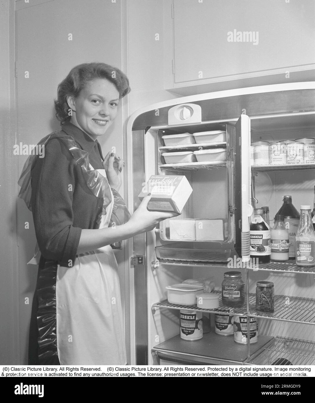 Dans la cuisine 1950s. Intérieur d'une cuisine et une jeune femme debout au réfrigérateur Leonard avec sa porte ouverte, montrant les boîtes et les bouteilles de différents aliments debout sur les étagères. Une armoire de congélation séparée est intégrée dans le réfrigérateur, une fonctionnalité assez nouvelle à ce moment. Elle porte un tablier en plastique. Elle est Haide Göransson, 1928-2008, mannequin et actrice. Suède 1950 Kristoffersson ref AU23-2 Banque D'Images