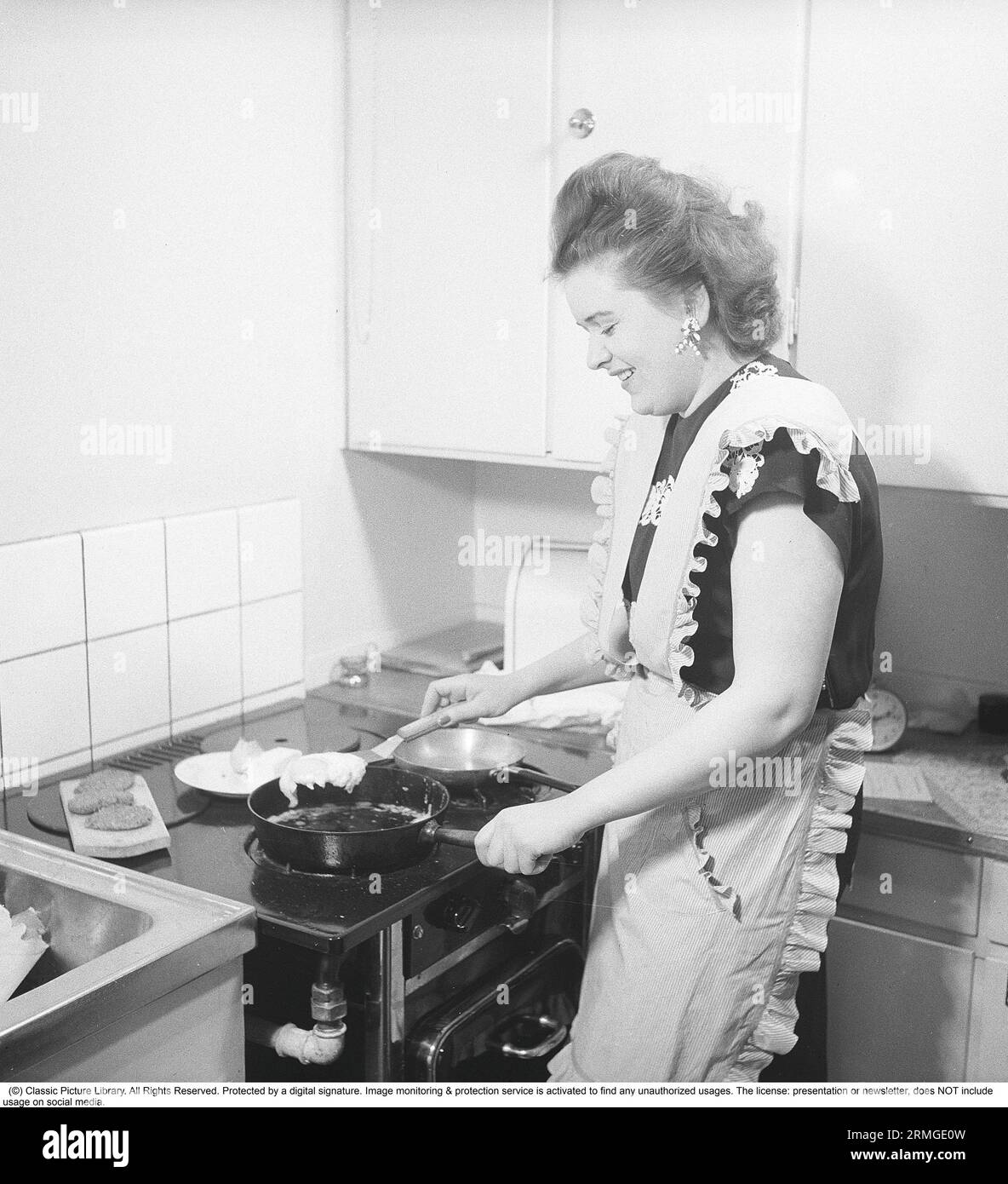 Dans la cuisine 1940s. Intérieur d'une cuisine et une jeune femme debout à la cuisinière de cuisine frire des œufs dans la poêle. Elle porte un tablier. Suède 1947. Kristoffersson réf. AB3-2 Banque D'Images