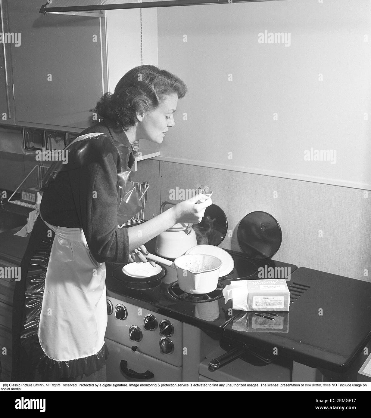 Dans la cuisine 1950s. Intérieur d'une cuisine et une jeune femme debout à la cuisinière de cuisine avec quelque chose de cuisson qui est dégustation. Elle porte un tablier en plastique. Elle est Haide Göransson, 1928-2008, mannequin et actrice. Suède 1950 Kristoffersson ref AU23-1 Banque D'Images