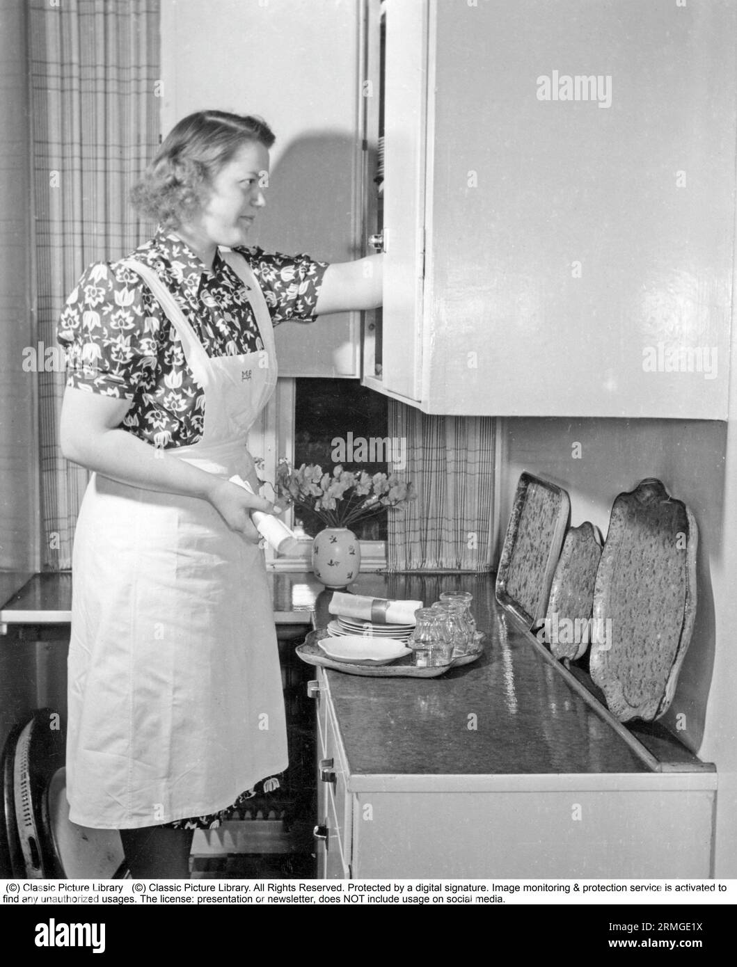 maison des années 1950. Une femme est vue préparer la table pour le dîner et dispose les assiettes et les verres qu'elle prend d'une armoire sur un plateau Suède 1950 Banque D'Images