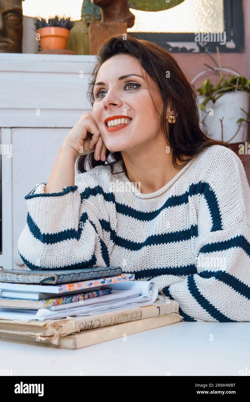 image verticale heureuse inspirée femme caucasienne française assise à la maison appuyée sur des livres à la table regardant vers le haut souriant et pensant. Banque D'Images