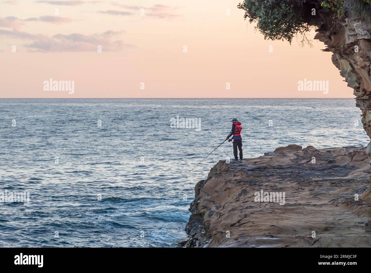 Un homme portant un gilet de sauvetage se tient debout à pêcher dans les rochers à l'extrémité sud populaire d'Avoca Beach sur la côte centrale de la Nouvelle-Galles du Sud, en Australie Banque D'Images
