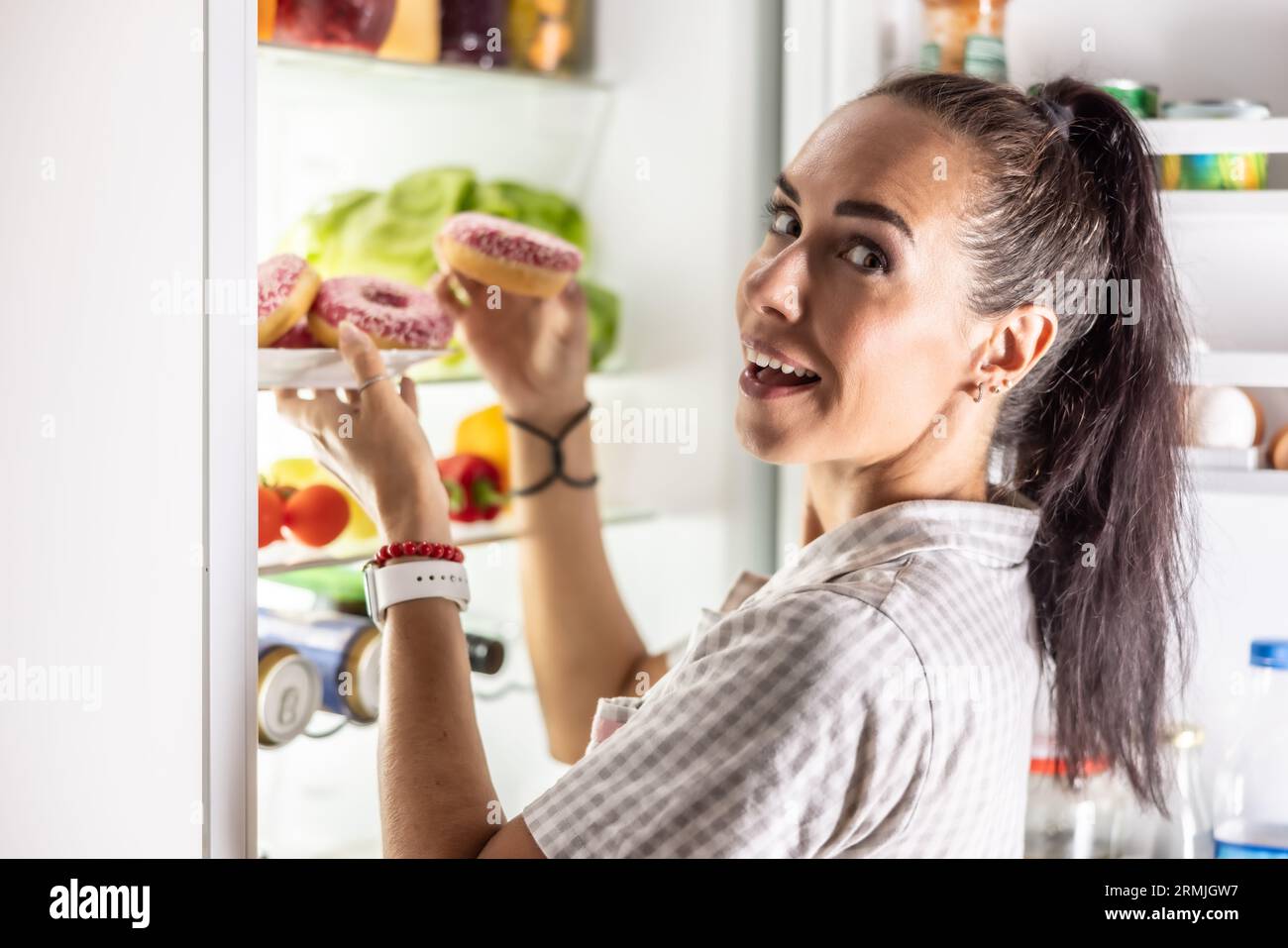 Une jeune femme affamée est sur le point de manger des beignets sucrés par le réfrigérateur ouvert tard dans la soirée. Banque D'Images
