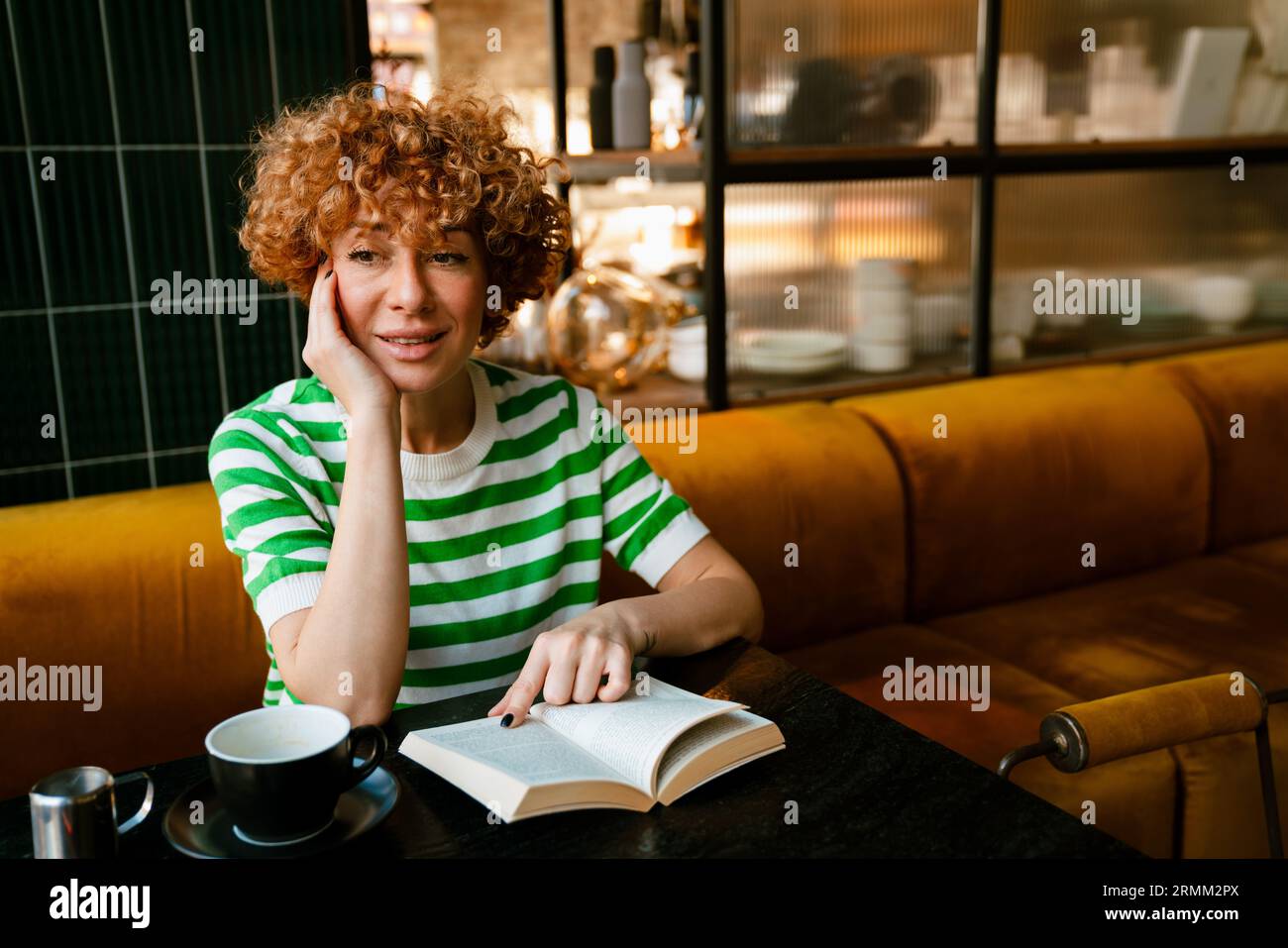 Femme blanche moyenne avec les cheveux bouclés lisant le livre tout en étant assis par la table dans le café Banque D'Images