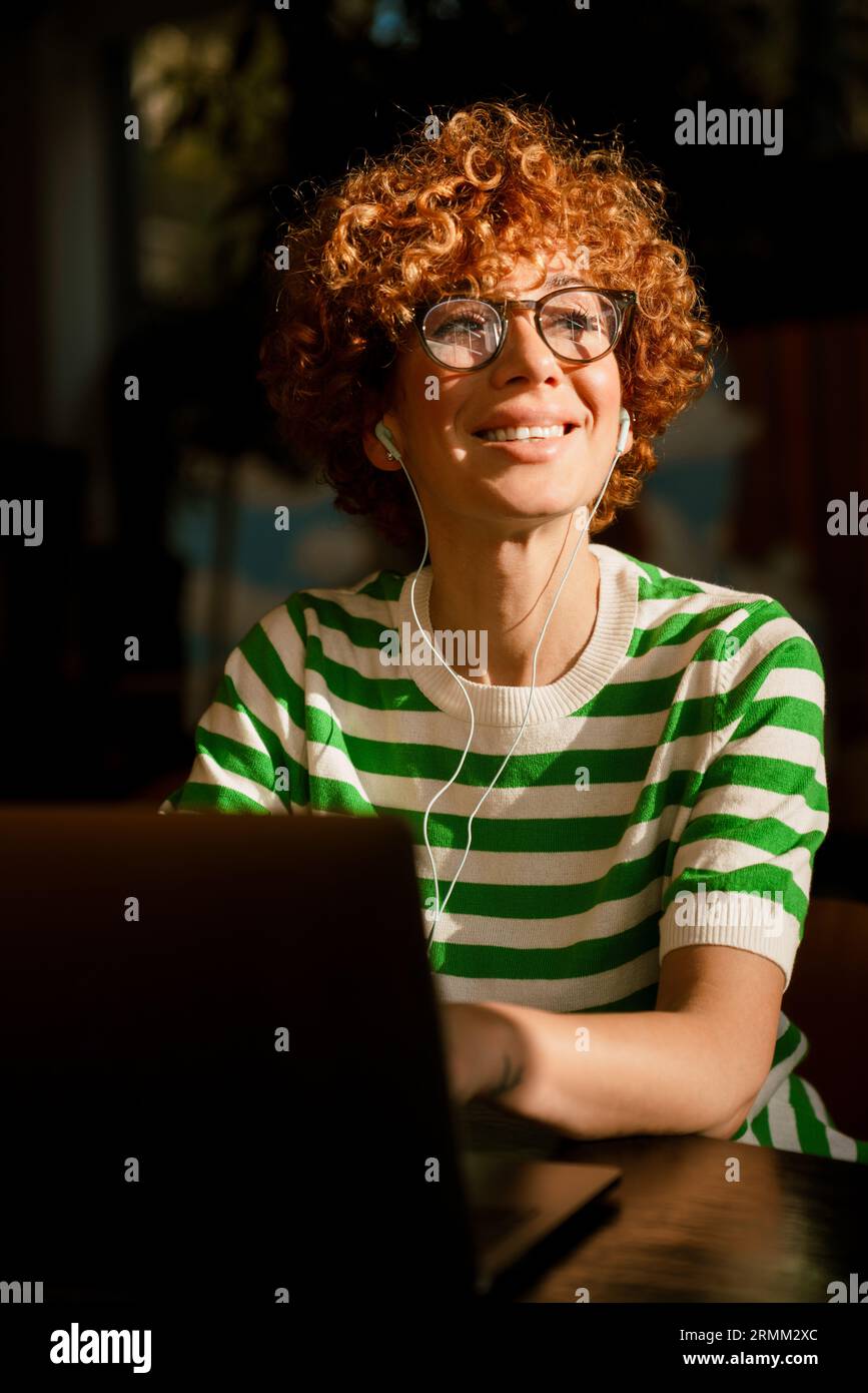 Souriant gingembre d'âge moyen dans des écouteurs et en utilisant un ordinateur portable tout en étant assis dans un café à l'intérieur Banque D'Images