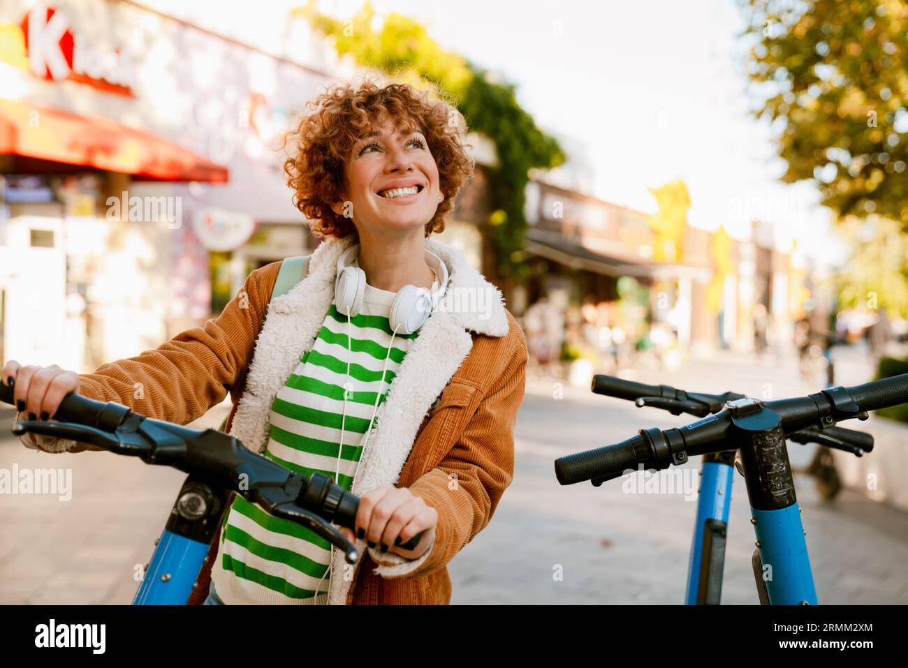 Gingembre souriant femme d'âge moyen avec scooter électrique et écouteurs sur son cou debout à l'extérieur dans la rue de la ville Banque D'Images