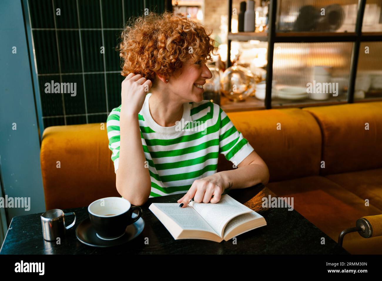 Gingembre gai femme d'âge moyen portant un t-shirt rayé lisant un livre et buvant du café tout en étant assis dans un café à l'intérieur Banque D'Images