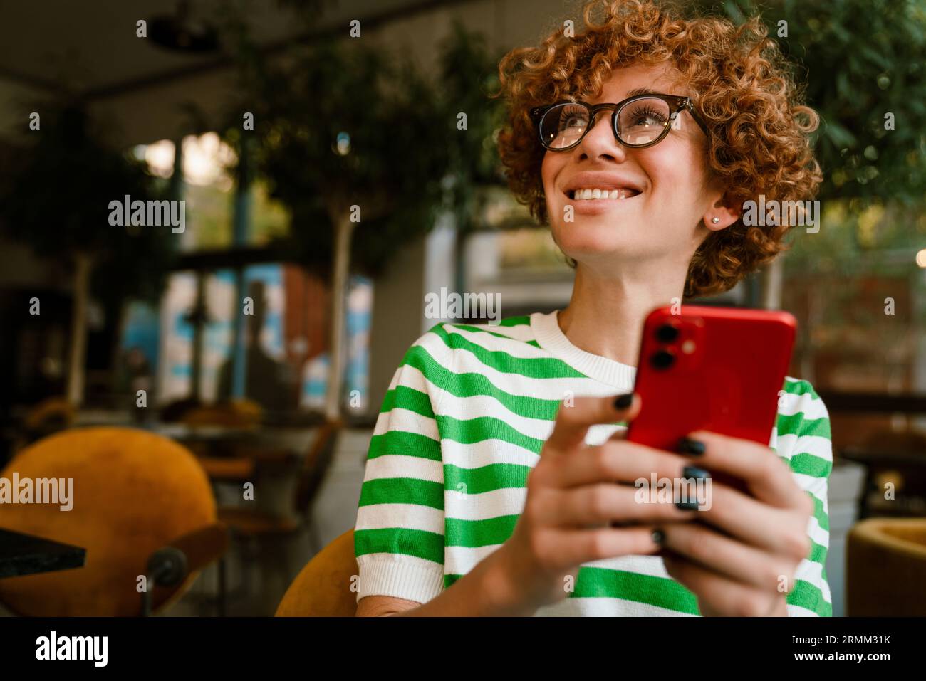 Femme blanche moyenne avec les cheveux bouclés souriant et utilisant le téléphone portable tout en étant assis dans le café Banque D'Images