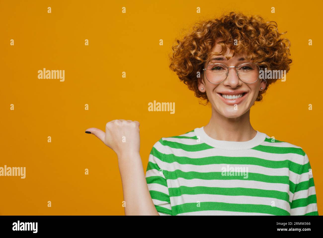 Femme bouclé de gingembre dans des lunettes souriant et pointant le doigt de côté isolé sur fond jaune Banque D'Images