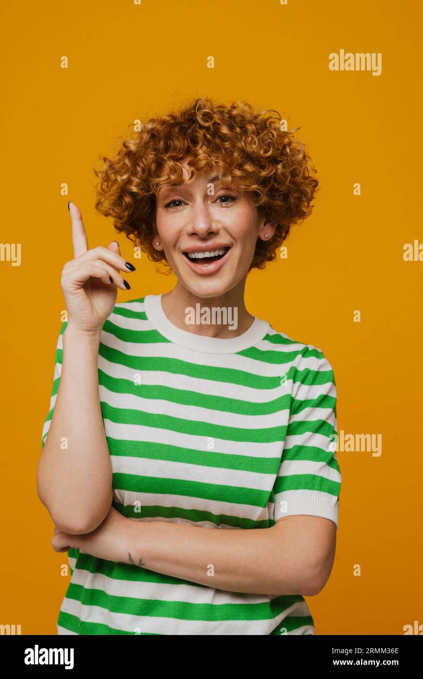 Femme de gingembre en t-shirt rayé souriant et pointant le doigt vers le haut isolé sur fond jaune Banque D'Images