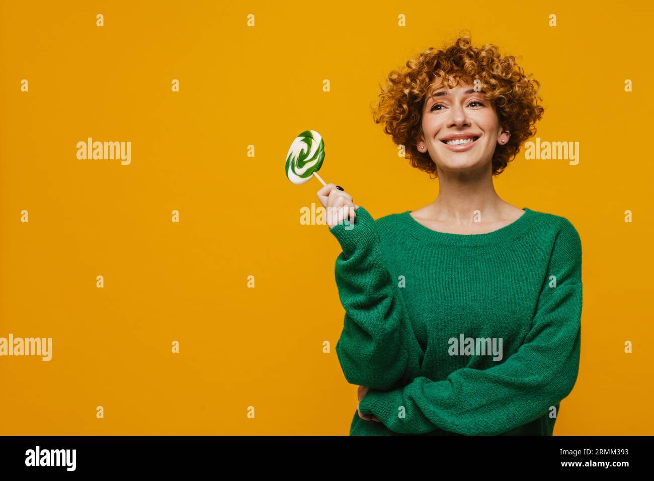 Femme de gingembre frisé souriant tout en posant avec sucette isolé sur fond jaune Banque D'Images