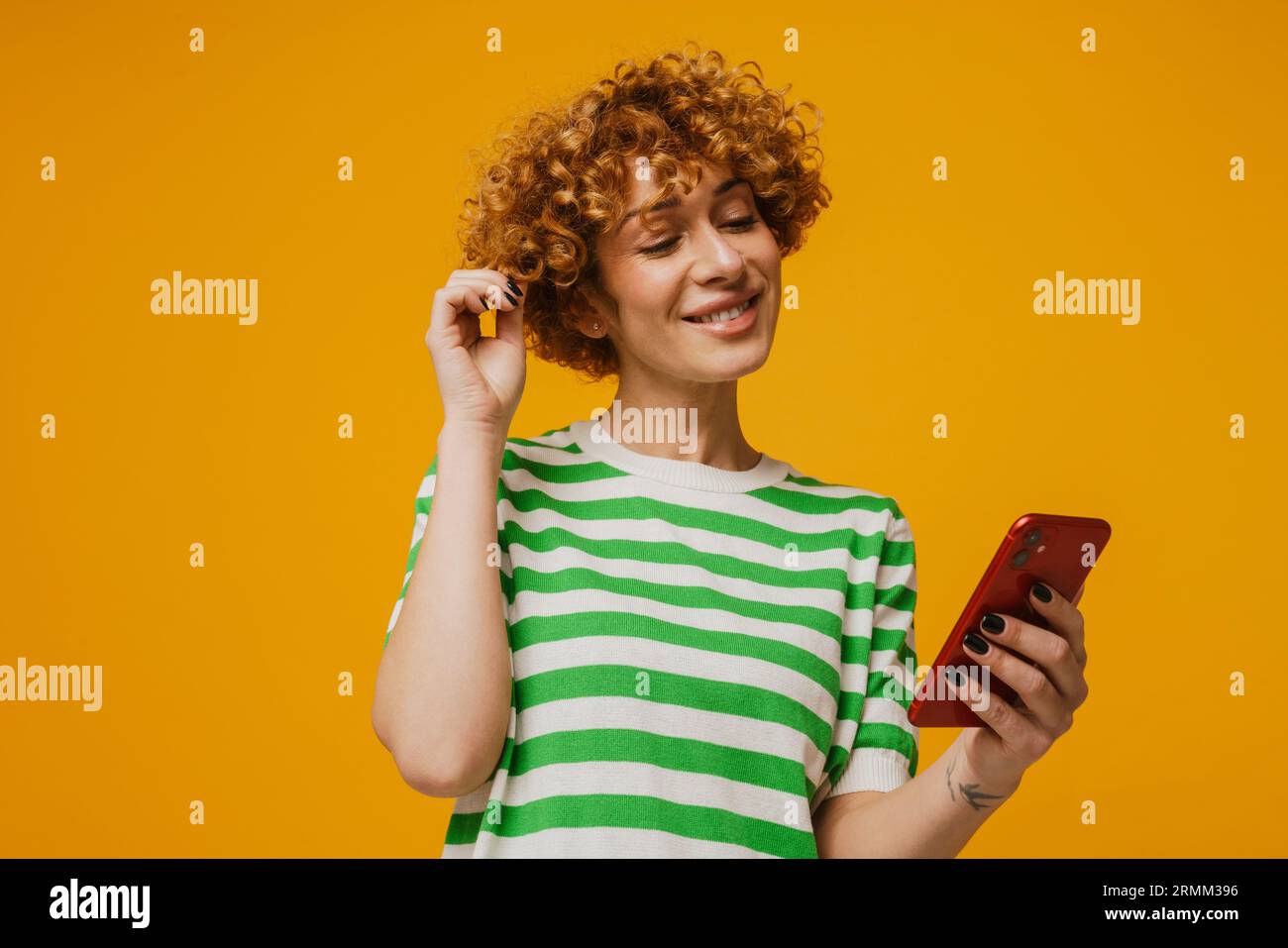 Femme bouclé de gingembre en t-shirt rayé souriant et en utilisant un téléphone portable isolé sur fond jaune Banque D'Images
