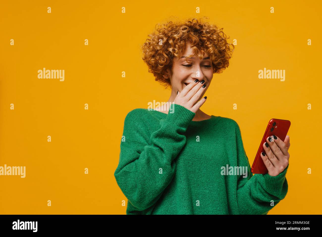 Femme mature de gingembre utilisant le téléphone portable et couvrant sa bouche avec la main isolée sur le mur jaune Banque D'Images