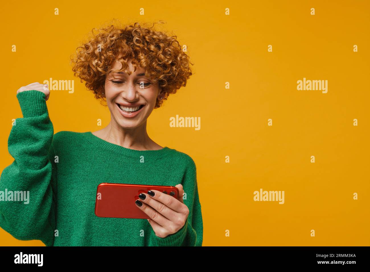 Femme bouclé de gingembre faisant le geste gagnant et utilisant le téléphone portable isolé sur fond jaune Banque D'Images
