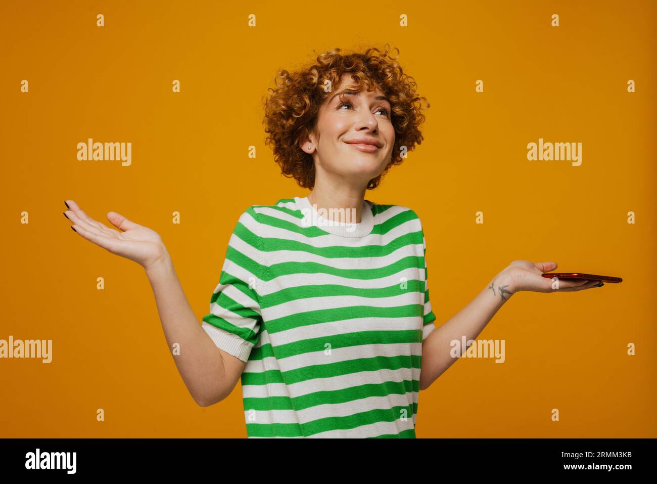 Femme bouclé de gingembre dans un t-shirt rayé gestant et utilisant un téléphone portable isolé sur fond jaune Banque D'Images