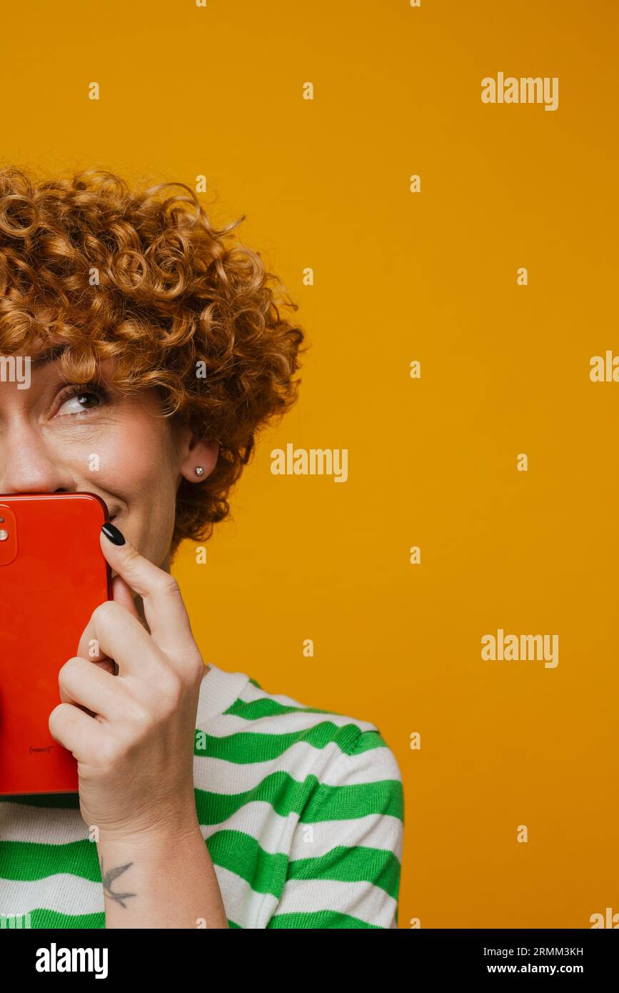 Femme de gingembre en t-shirt rayé souriant et posant avec téléphone portable isolé sur fond jaune Banque D'Images